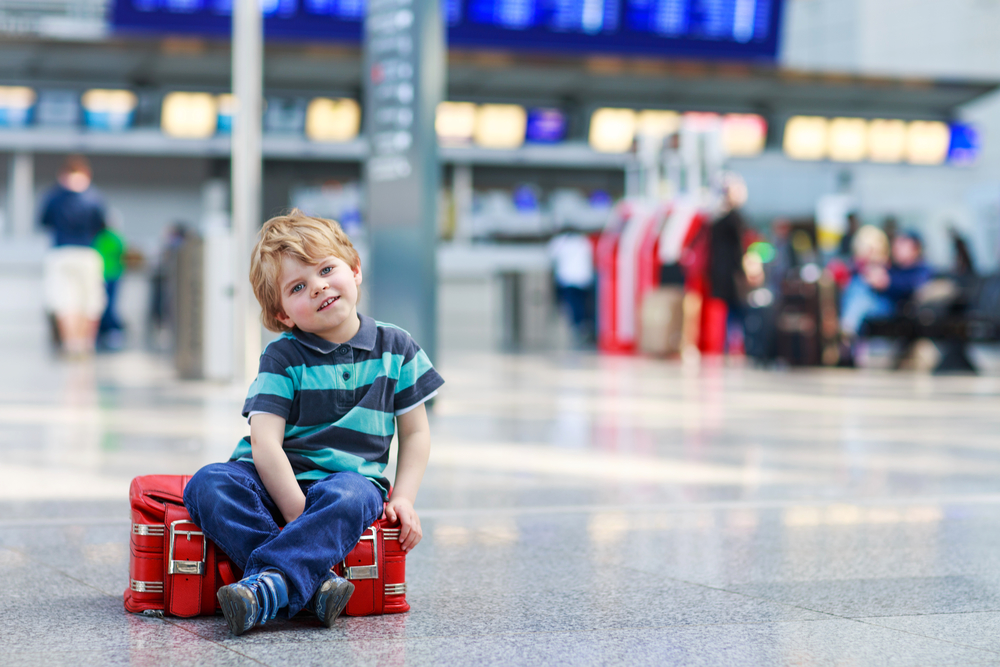 Blond boy of 2 years sitting on suitcase at the airport, indoors and waiting for going on vacations.