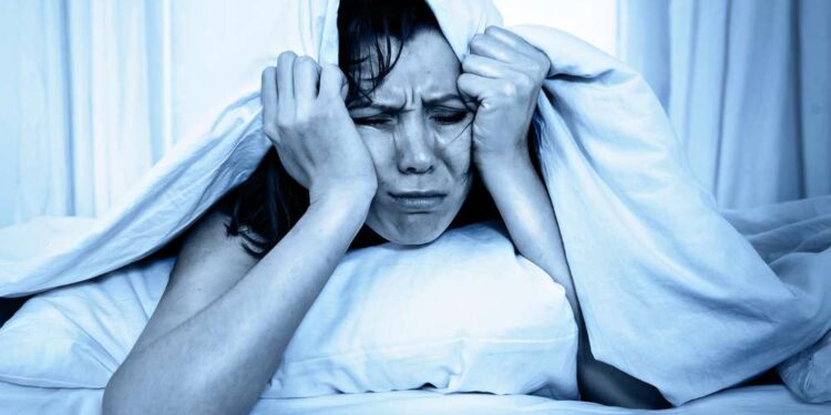 young moody woman in bed suffering stress with her insomnia problem and sleeping disorder or hangover covering with blanket and bedclothes on studio blue lighting