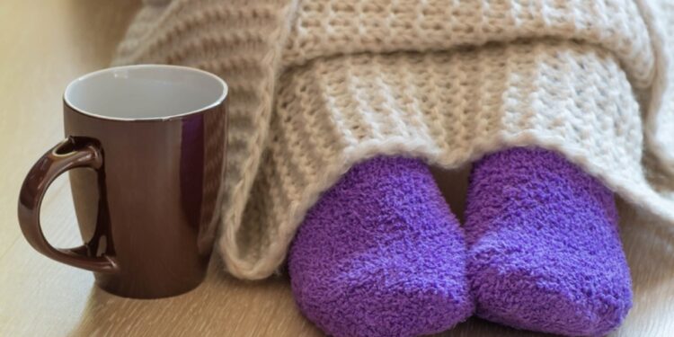 Cold concept with teacup near the legs which are covered with fluffy warm blanket and wearing fluffy warm purple socks