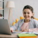 happy child looking at camera while sitting at desk and using laptop at home